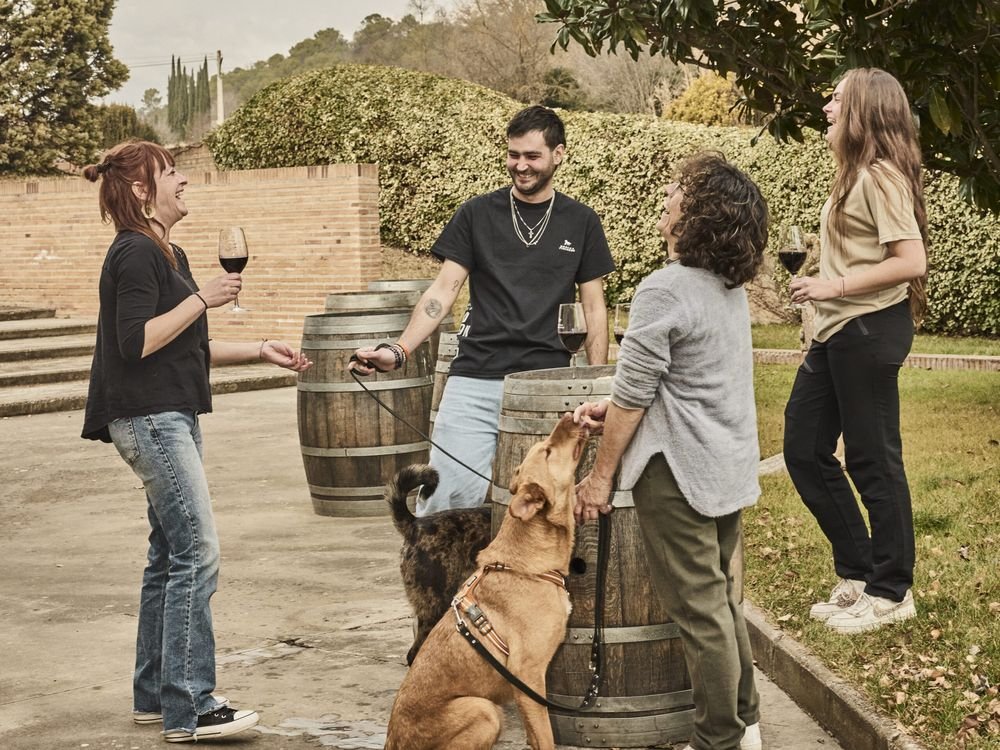Un perro pide caricias a un grupo que cata vino junto a unas barricas